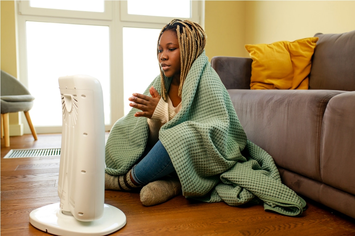 african american woman in blue blanket warming near electric heater at home.